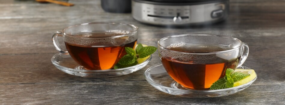 Two cups of tea on saucers, each with a slice of lemon and some mint leaves. Behind is the bottom of a KitchenAid Electric Kettle, the name "KitchenAid" is visible.